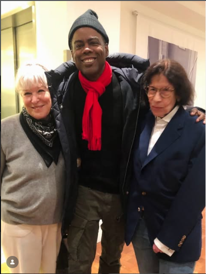 Three people smiling indoors: a man with a red scarf between two women in light-colored outfits, all posing together.