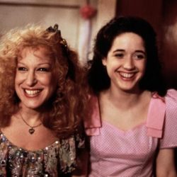 Two smiling women posing together indoors, one with curly blonde hair wearing a sequined top and the other with dark hair in a pink dress on the right.