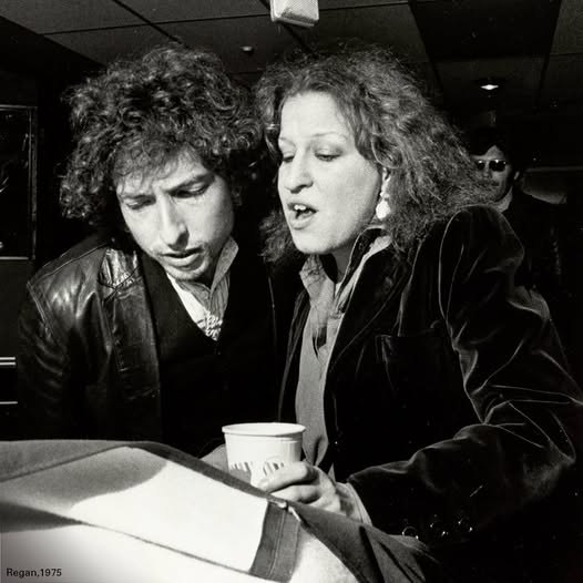 Two men with curly hair lean over a car hood, one holding a paper cup, examining something in a black?and?white 1970s scene.
