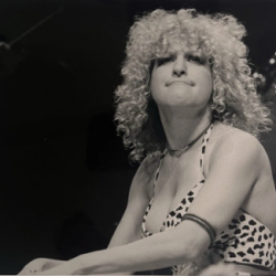 Black-and-white photo of a female singer with curly hair on stage, wearing a leopard-print top and a necklace, looking upward with a confident expression.