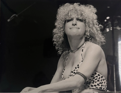 Black-and-white photo of a female singer with curly hair on stage, wearing a leopard-print top and a necklace, looking upward with a confident expression.