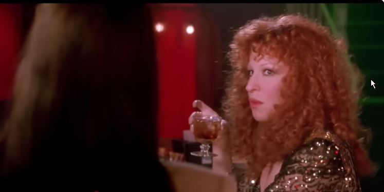 Red-haired woman in a sequined dress holding a cocktail glass at a dimly lit bar.