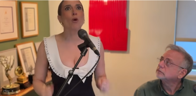 Woman singing into a microphone while an older man watches in a room with framed certificates on the wall and trophies on a shelf.