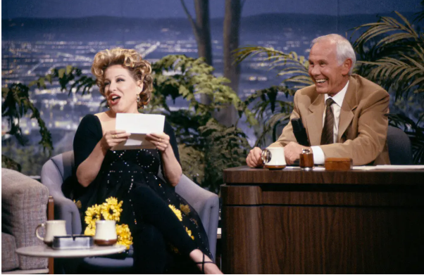 2026-05-05_11-52-00 - bootleg betty Female guest reads cue card while smiling on a TV talk show set; host sits at a wooden desk smiling beside her. A plants-filled backdrop and mugs on the desk.