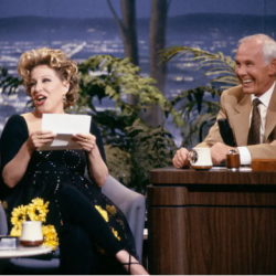 Talk-show scene with a female guest in a black dress showing a card while smiling, seated next to a smiling male host at a wooden desk with coffee mugs and plants behind them