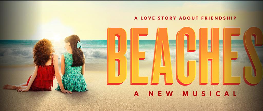 Two girls in red and turquoise dresses sit on a sunlit beach, with a large orange 'BEACHES' title and the subtitle 'A love story about friendship' and 'A NEW MUSICAL'.