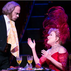 Two actors at a stage dinner: a man in a dark suit and gold waistcoat stands beside a table, while a woman in a red lace dress with a tall pink feathered hat gestures animatedly across it.