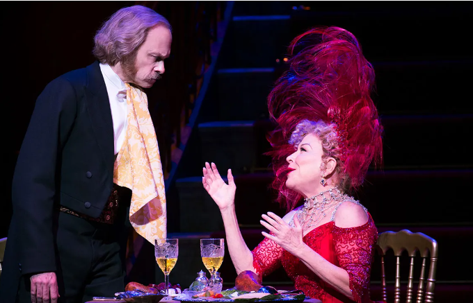 Two actors at a stage dinner: a man in a dark suit and gold waistcoat stands beside a table, while a woman in a red lace dress with a tall pink feathered hat gestures animatedly across it.