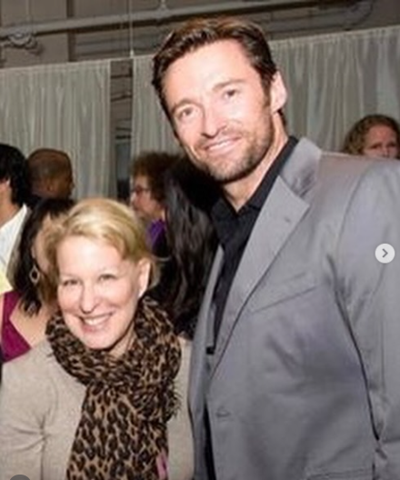 Smiling blonde woman in a beige top and leopard scarf, standing with a tall man in a light gray suit at an event.