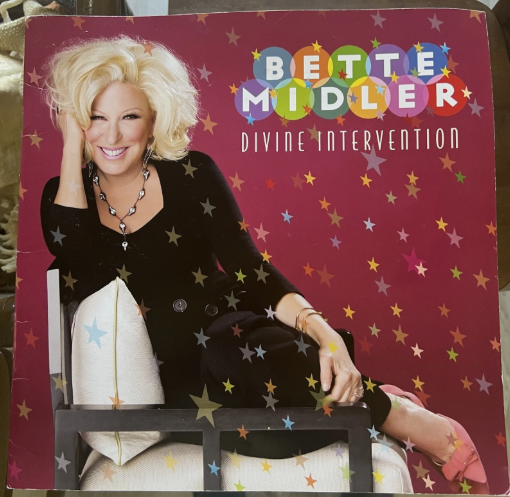 Bette Midler smiling on a pink star-covered album cover for 'Divine Intervention'.