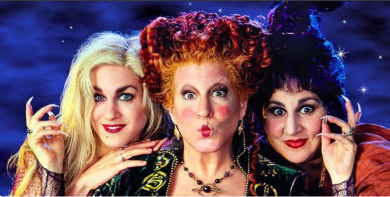 Three women in colorful witch costumes pose together against a starry night backdrop.
