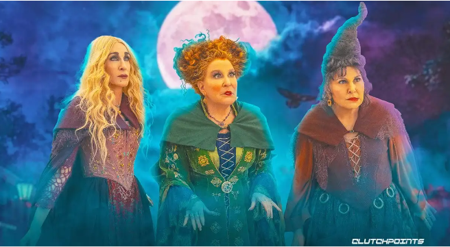 Three witches in colorful medieval costumes stand under a full moon in a blue-purple forest scene, looking ahead with serious expressions.