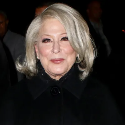 Older woman with silver hair wearing a black coat at a formal evening event, facing the camera with a slight smile.