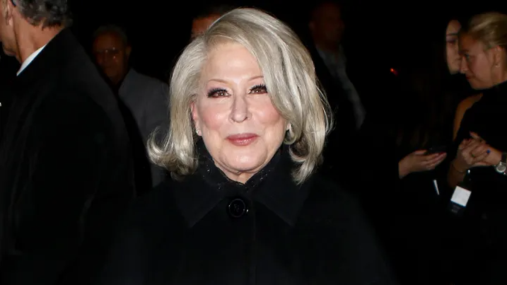 Older woman with silver hair wearing a black coat at a formal evening event, facing the camera with a slight smile.