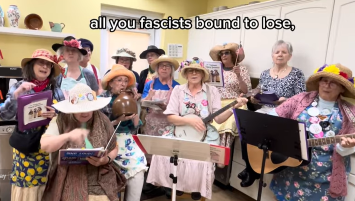 Group of elderly women wearing colorful hats, singing and playing instruments in a bright kitchen; caption reads 'all you fascists bound to lose'.