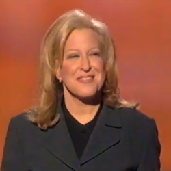 Smiling blonde woman in a dark blazer on a stage with an orange background.