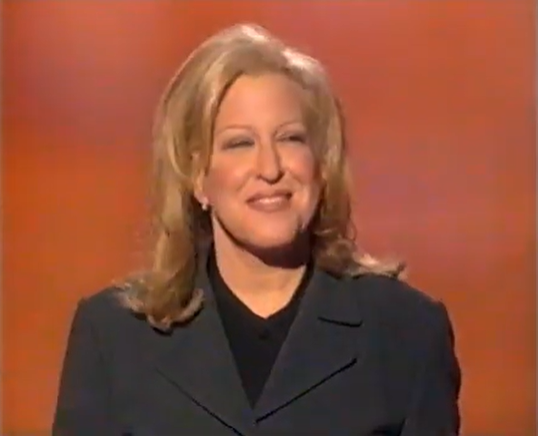 Smiling blonde woman in a dark blazer on a stage with an orange background.