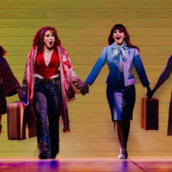 Group of women holding hands on stage, mid-performance, colorful retro outfits and suitcases in hand against a yellow backdrop.