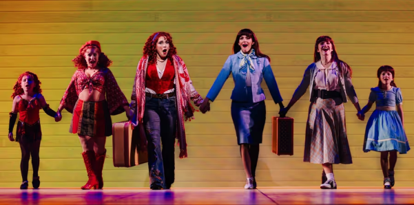 Group of women holding hands on stage, mid-performance, colorful retro outfits and suitcases in hand against a yellow backdrop.