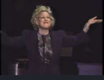 Woman with curly blonde hair on a dimly lit stage, arms outstretched and smiling.