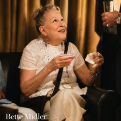 Older woman in a white lace blouse sits with a pink cocktail, as a man offers a drink nearby in a dim, gold-curtained room.