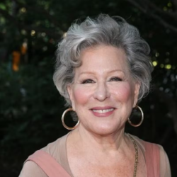 Smiling Bette Midler with short gray hair, wearing hoop earrings and a pink top, outdoors at dusk.