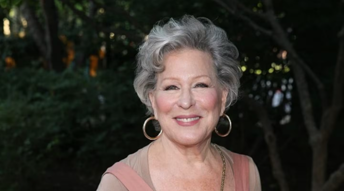 2026-05-30_07-25-38 - bootleg betty Smiling Bette Midler with short gray hair, wearing hoop earrings and a pink top, outdoors at dusk.