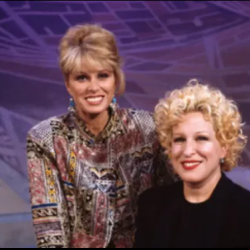 Two smiling women , Bette Midler and Joanna Lumley posing together in a studio with a blue geometric backdrop, one wearing a patterned blouse and the other in a black outfit.]