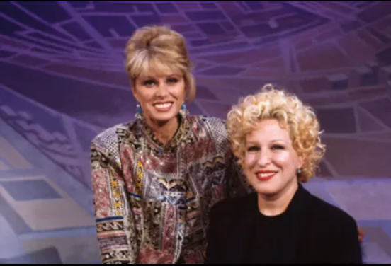 Two smiling women , Bette Midler and Joanna Lumley posing together in a studio with a blue geometric backdrop, one wearing a patterned blouse and the other in a black outfit.]