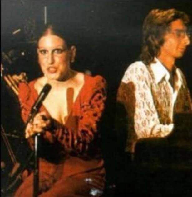 Female singer in a red outfit performs on stage into a microphone, with a male pianist in a patterned shirt seated at a piano beside her.