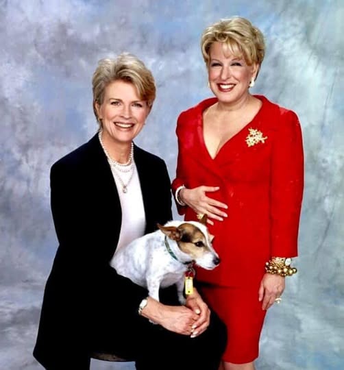 Two smiling women posing for a portrait; one in a black blazer holding a small dog wearing a collar, the other in a red dress with jewelry.