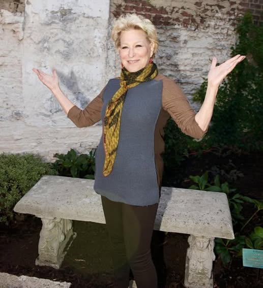 Bette Midler standing outdoors with arms raised, wearing a blue-gray tunic, brown sleeves, black pants, and a yellow scarf near a stone bench and brick wall.