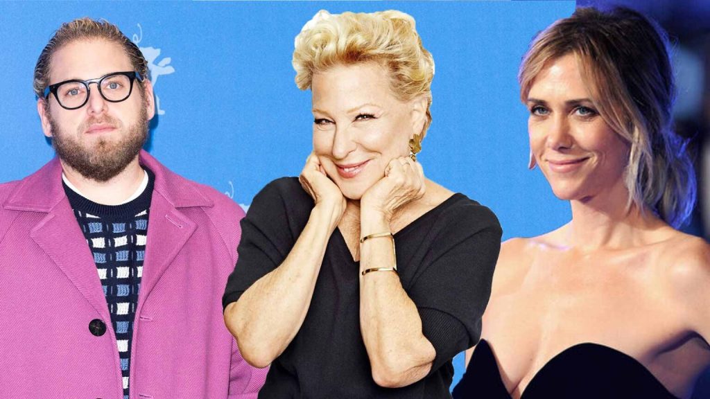 Three adults pose for a photo against a blue backdrop: a man in a pink jacket and glasses on the left, a smiling blonde woman center with hands on her cheeks, and a woman in a black strapless dress on the right.