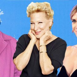 Three adults pose for a photo against a blue backdrop: a man in a pink jacket and glasses on the left, a smiling blonde woman center with hands on her cheeks, and a woman in a black strapless dress on the right.