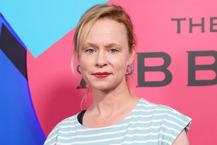 Woman with blonde hair and hoop earrings poses for photos in a light blue striped top against a pink and blue backdrop at an event.