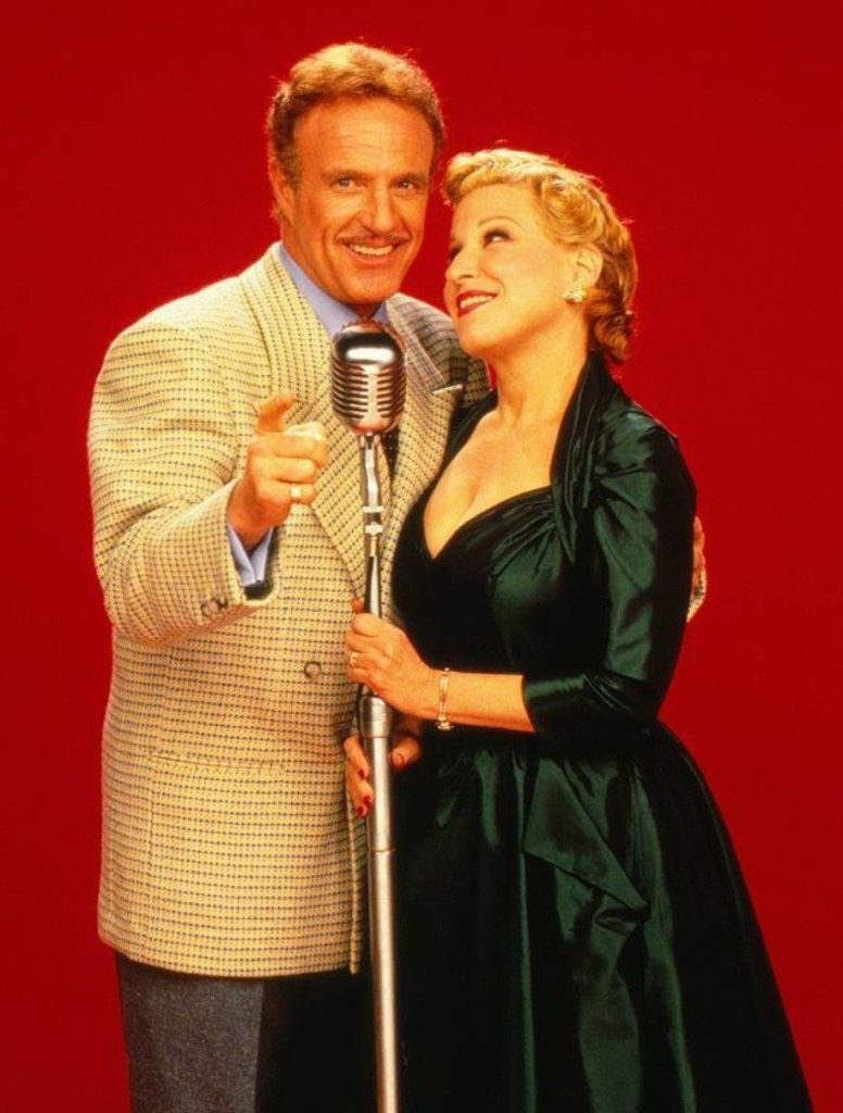 A man in a beige checkered blazer and a woman in a dark green gown stand close together, singing into a vintage microphone against a red backdrop.