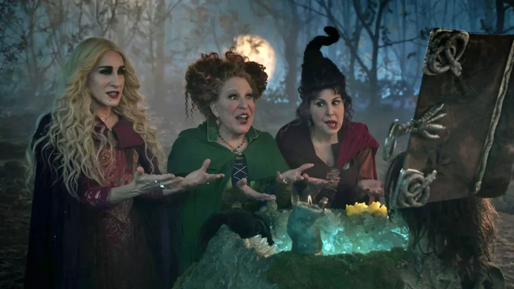 Hocuspocus2-sandersons - bootleg betty Three witches in a moonlit forest lean over a glowing cauldron, gesturing as they cast a spell.