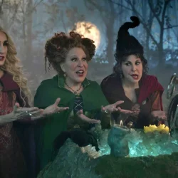 Three witches in a moonlit forest lean over a glowing cauldron, gesturing as they cast a spell.