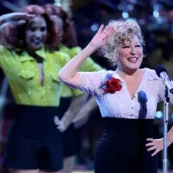 Bette Midler waves at a microphone on stage with backup dancers in bright outfits behind her, during a live performance.