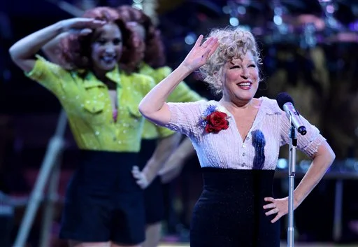 Bette Midler waves at a microphone on stage with backup dancers in bright outfits behind her, during a live performance.