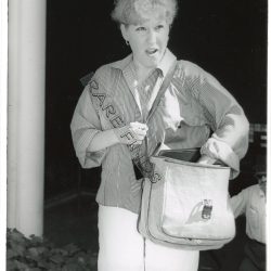 Black-and-white photo of a woman with short light hair wearing a striped button-down shirt, carrying a tote-style bag and papers, standing near a doorway outdoors.