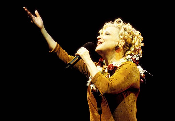 Female singer on stage, microphone in hand, arm raised in a dramatic gesture against a black background.
