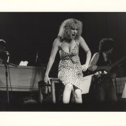 Female singer in a leopard-print dress belts into a mic on stage, flanked by a pianist and guitarist in a live performance.