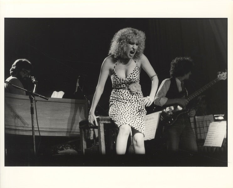 Female singer in a leopard-print dress belts into a mic on stage, flanked by a pianist and guitarist in a live performance.