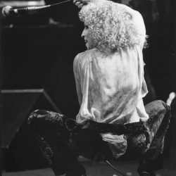 Back view of a curly-haired performer kneeling on stage, holding a microphone cable overhead.