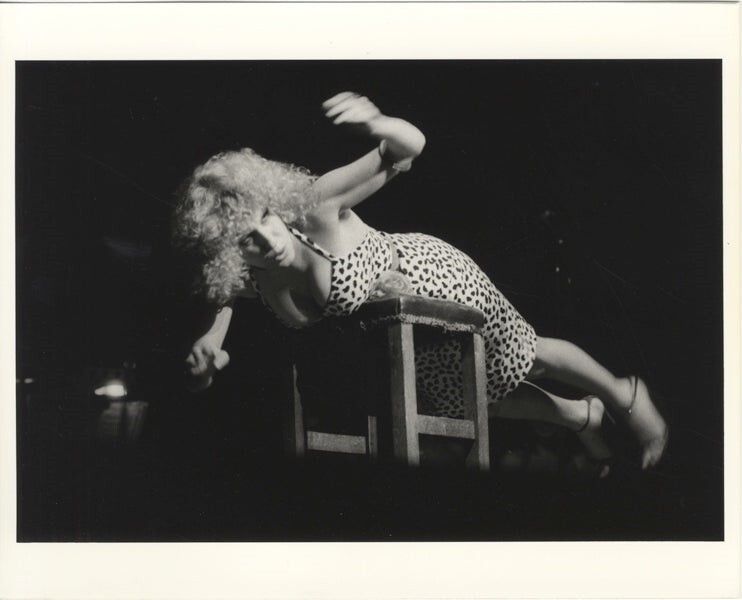 Black-and-white photo of a performer in a leopard-print outfit leaning over a wooden chair, arms raised, mid-dance on a dimly lit stage.
