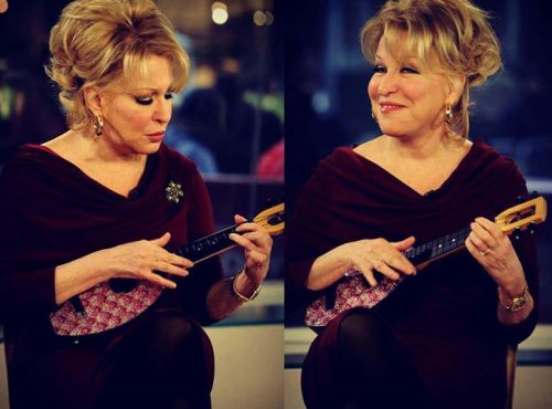 Older woman in a burgundy dress playing a small string instrument (ukulele) on a TV studio set; two mirrored poses show different facial expressions.