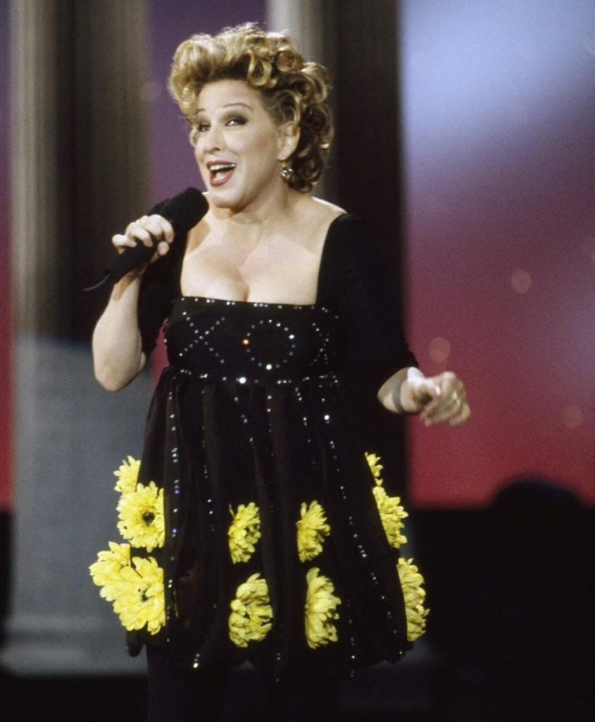 Female singer on stage, wearing a black sequined dress with yellow flower embroidery, holding a microphone and singing.