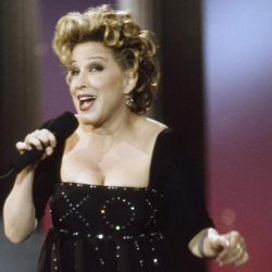 Female singer on stage, wearing a black sequined dress with yellow flower embroidery, holding a microphone and singing.