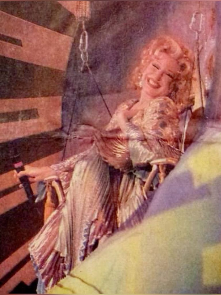 Smiling woman in a patterned dress sitting against a striped wooden wall.